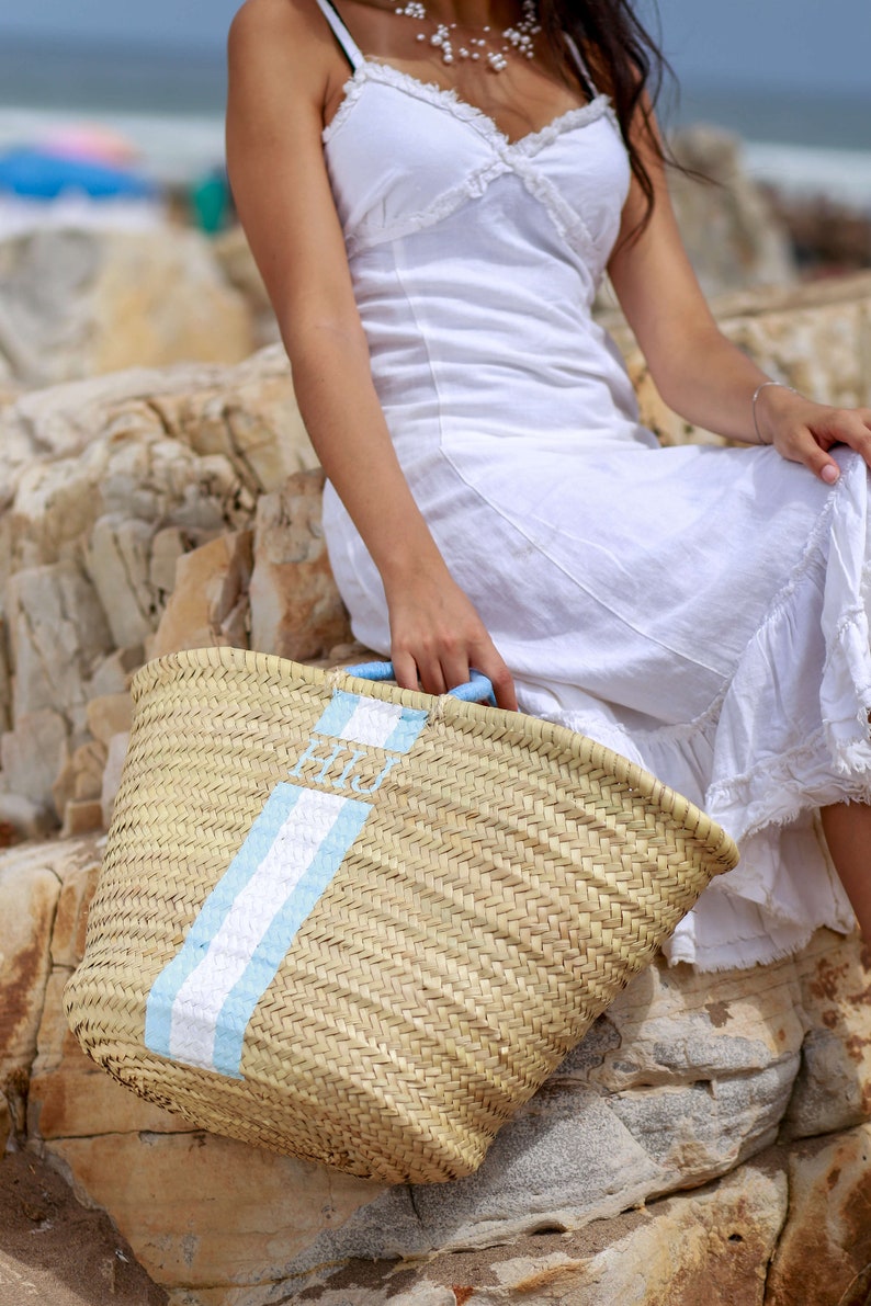 May include: A large, woven straw tote bag with a blue and white stripe and the letters "HIJ" printed on the front. The bag is sitting on a rocky surface.