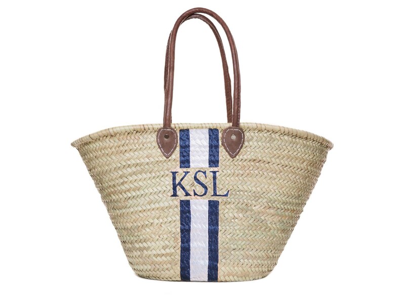 May include: Woven straw tote bag with brown leather handles and accents. Features a navy and white stripe design with embroidered initials "KSL". A stylish and practical accessory for carrying items.