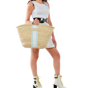May include: A large, natural woven basket with a blue and white striped handle and a blue leather strap. The basket is filled with a variety of items, including a white dress, a pair of white boots, and a black belt.