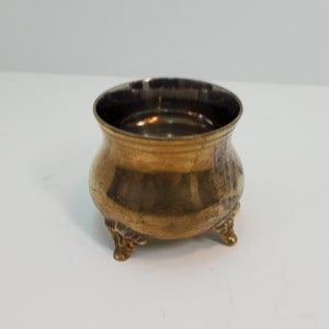 Puede incluir: Una pequeña olla de metal de color dorado con tres patas. La olla tiene una textura martillada y una forma redonda.