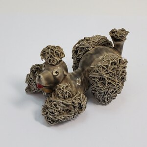 May include: A ceramic figurine of a brown poodle lying down with its head turned to the side. The poodle has a red bow around its neck.