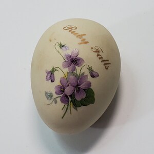 Könnte beinhalten: Ein weißer, eiförmiger Keramik-Anhänger mit einem violetten Blumendesign und dem Text "Rugby Falls" in goldenen Buchstaben.