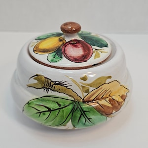 May include: A white ceramic jar with a lid, decorated with a hand-painted design of green leaves, brown branches, and red and yellow fruit. The lid has a brown knob.