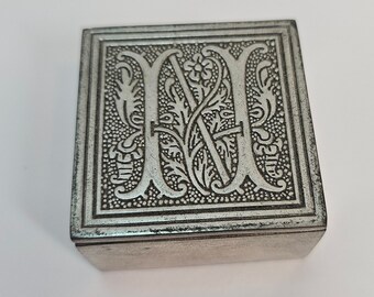 May include: A silver metal box with an ornate design featuring the letter "M" in a decorative font.