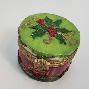 May include: A small, round decorative box with a hinged lid. The lid is green with a holly and berry design. The box's base is burgundy with gold accents and the year "2001" in gold. A festive holiday-themed trinket box.