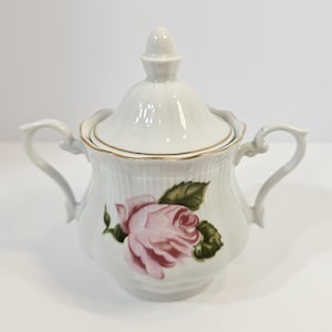 May include: A white porcelain sugar bowl with a lid, featuring a delicate pink rose floral pattern with green leaves on the front. The bowl has two curved handles, a ribbed texture, and a gold-trimmed rim on both the lid and the bowl body.