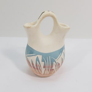 Könnte beinhalten: Eine kleine, weiße Keramikvase im indianischen Stil mit Henkel. Die Vase hat ein blaues und orangefarbenes Design mit weißen Akzenten.