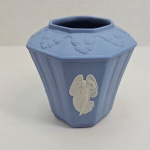 May include: A small, light blue ceramic vase with a white angel design. The vase has a hexagonal shape and a scalloped rim.