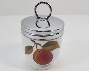 Könnte beinhalten: Weißer Eierbecher aus Keramik mit silberfarbenem Deckel und Griff. Der Becher hat ein gemaltes Design einer Pflaume mit grünen Blättern. Der Deckel und der Griff sind glänzend und reflektierend.