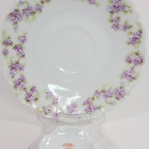 Puede incluir: Una taza de té y un platillo de porcelana blanca con un patrón floral. El platillo tiene un borde festoneado y la taza tiene un asa dorada. La parte inferior de la taza tiene una marca que dice "Elite Limoges France".