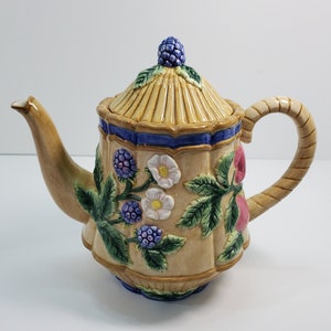 May include: A ceramic teapot with a floral design featuring blackberries, white flowers, and green leaves. The teapot has a beige base with blue and green accents. The lid is shaped like a dome and has a brown and beige pattern.