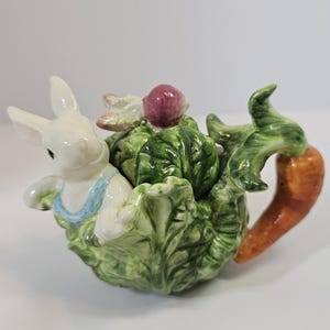Może przedstawiać: Ceramiczny czajnik w kształcie kapusty z białym królikiem w niebieskim śliniaczku. Czajnik ma uchwyt w kształcie marchewki i mały czerwony owoc na górze. Kapusta jest zielona z białymi akcentami.