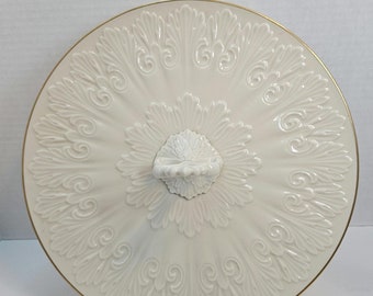 Könnte beinhalten: Eine weiße Keramik-Servierplatte mit goldfarbenem Rand und einem verzierten Design. Die Platte hat einen Griff in der Mitte und steht auf einem schwarzen Ständer.