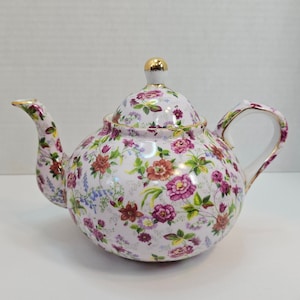 May include: A white porcelain teapot with a floral pattern in shades of pink, red, and green. The teapot has a gold trim and a lid with a gold knob.