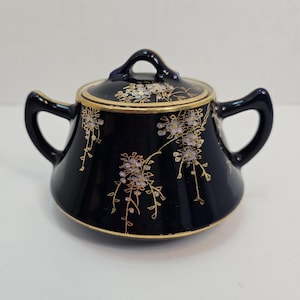 May include: A small, dark blue ceramic sugar bowl with a lid. The bowl is decorated with gold trim and a floral pattern in white and gold.
