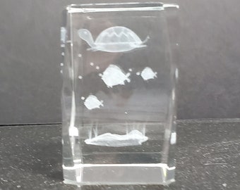 May include: A clear glass paperweight with an etched design of a turtle swimming with fish. The paperweight is rectangular and has a bevelled edge.