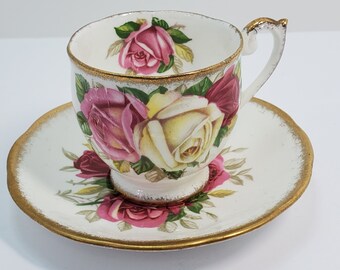 May include: A white teacup and saucer set with a floral design featuring pink, yellow, and red roses. The set has a gold trim around the edges.
