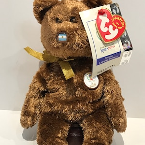 May include: Brown plush teddy bear wearing a gold ribbon and a blue and white pin with the flag of Argentina. The bear is sitting on a wooden base and has a Ty Beanie Baby tag attached to its right ear. The tag reads "Champion the Beanie Baby" and "2002 FIFA World Cup Korea/Japan Official Licensed Product H015589986".