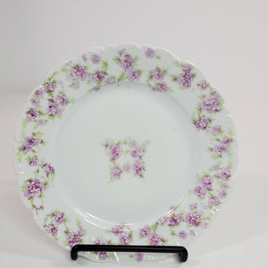 Puede incluir: Un plato de porcelana blanca con un borde festoneado y un diseño floral. El plato está decorado con pequeñas flores moradas y hojas verdes.
