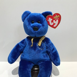 May include: Blue Ty Beanie Baby bear with a gold ribbon and a star-shaped patch on its chest. The bear is sitting on a brown stand. The Ty Beanie Baby tag is attached to the bear's right ear.