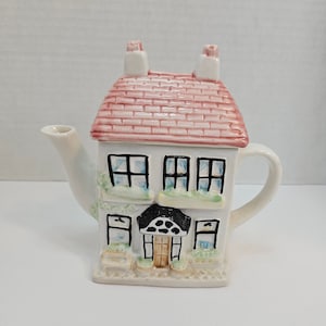 Peut inclure: Une théière en céramique blanche en forme de maison avec un toit rose et des fenêtres noires. La théière a un bec verseur et une poignée.