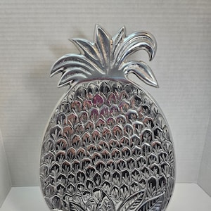 May include: A silver pineapple-shaped serving tray with a detailed design of the fruit's leaves and skin. The tray is on a black stand.