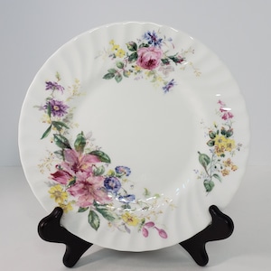 May include: A white porcelain plate with a floral design. The plate has a scalloped edge and features a variety of pink, purple, yellow, and blue flowers.