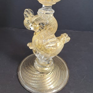 Puede incluir: Una copa de vidrio con pan de oro y base en forma de pájaro. La copa es de vidrio transparente con detalles en pan de oro. La base es un pájaro con acabado en pan de oro.