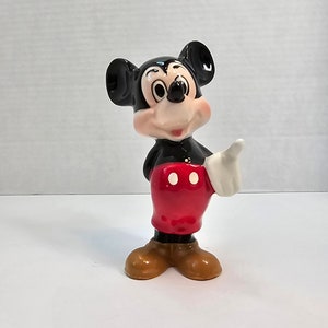 May include: A ceramic figurine of Mickey Mouse. He is standing with his right hand raised and his left hand on his hip. He is wearing his signature red shorts with two white buttons and yellow shoes.