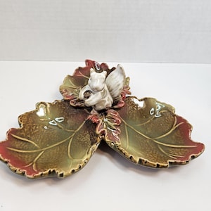 Puede incluir: Una fuente de cerámica con forma de tres hojas, con una figura de ardilla en el centro. La fuente tiene un acabado brillante con tonos verdes, marrones y rojos. La ardilla sostiene una nuez.