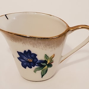 Pode incluir: Um creamer branco com uma borda dourada e um desenho de flor azul. O creamer tem uma alça e é feito de porcelana.