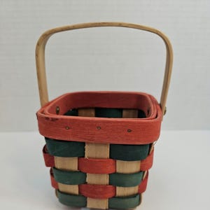 May include: A small, square-shaped basket with a woven design in red, green, and natural wood tones. The basket has a wooden handle and is about the size of a 25 pence coin.