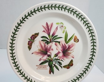 May include: A white ceramic plate with a green border featuring a botanical illustration of pink rhododendron flowers and butterflies. The text "RHODODENDRUM LILEIFLORUM Lily Flowered Azalea" is printed on the plate.