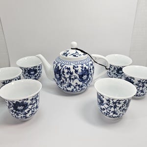 Peut inclure: Service à thé en porcelaine blanche avec un motif floral bleu. L'ensemble comprend une théière et six tasses assorties. La théière a un couvercle et un cordon noir. Les tasses sont petites et rondes.