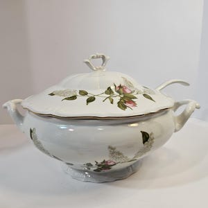 May include: A white porcelain soup tureen with a matching lid and a ladle. The tureen features a delicate floral pattern of pink roses and white lilac clusters, accented with gold trim along the edges and a decorative handle on the lid.