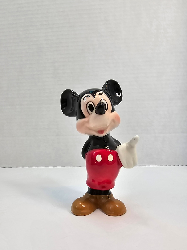 May include: A ceramic figurine of Mickey Mouse. He is standing with his right hand raised and his left hand on his hip. He is wearing his signature red shorts with two white buttons and yellow shoes.