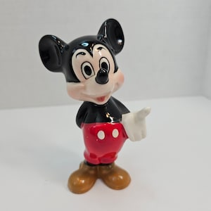 May include: A ceramic figurine of Mickey Mouse. He is standing with his right hand raised and pointing. He is wearing his signature red shorts with white polka dots, black shoes, and white gloves.