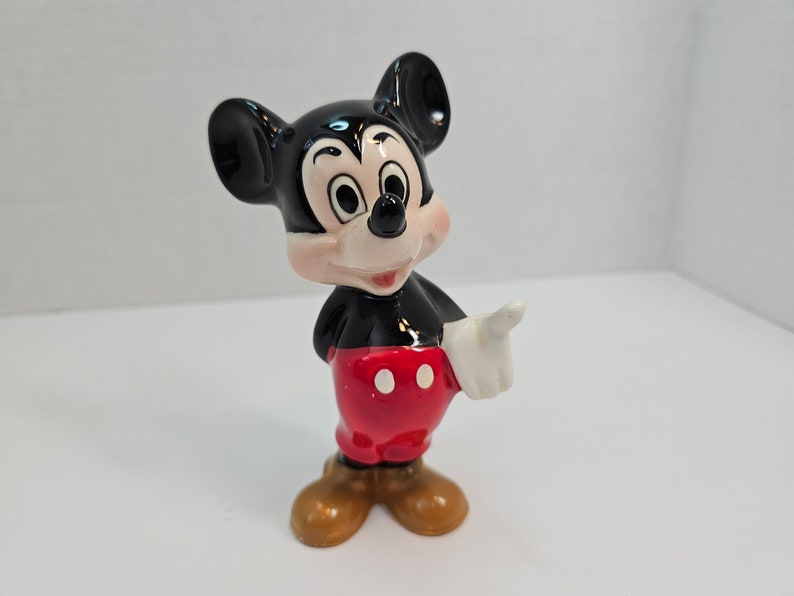 May include: A ceramic figurine of Mickey Mouse. He is standing with his right hand raised and pointing. He is wearing his signature red shorts with white polka dots, black shoes, and white gloves.