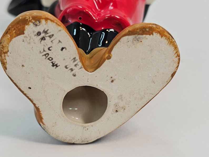 May include: The base of a ceramic figurine with a red face and black eyes. The figurine is marked "Walt Disney" and "Made in Japan".