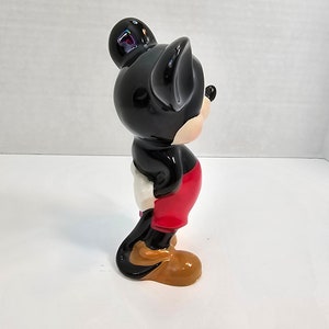 May include: A black and red ceramic figurine of Mickey Mouse. The figurine is standing with its right arm raised and its left arm at its side. The figurine is wearing red shorts and yellow shoes.