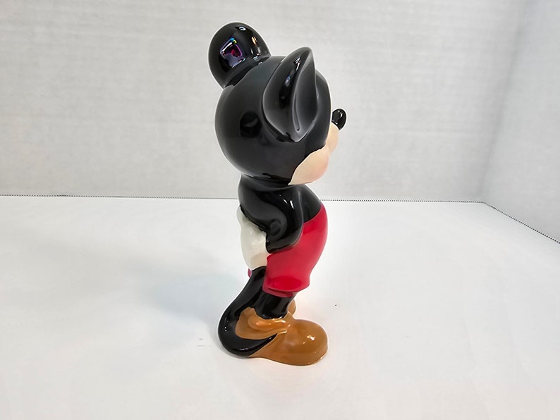 May include: A black and red ceramic figurine of Mickey Mouse. The figurine is standing with its right arm raised and its left arm at its side. The figurine is wearing red shorts and yellow shoes.