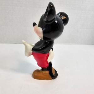 May include: A black and white ceramic figurine of Mickey Mouse. He is wearing red shorts and brown shoes. He is standing with his right leg raised and his left arm outstretched.