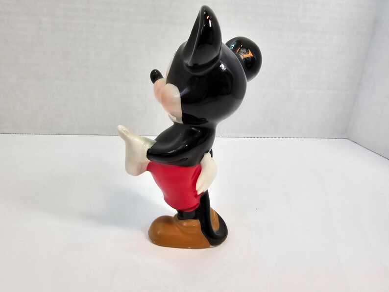 May include: A black and white ceramic figurine of Mickey Mouse. He is wearing red shorts and brown shoes. He is standing with his right leg raised and his left arm outstretched.