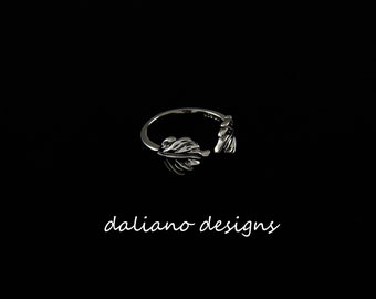 May include: A silver-colored ring with a leaf design. The ring has an open design with two leaf-shaped ends. The ring is set against a black background. The text "daliano designs" is written in a cursive font.