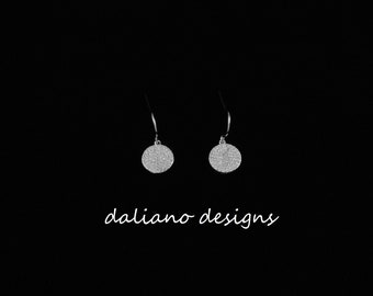 May include: A pair of silver earrings with a round, sparkly design. The earrings are hanging from a black background.