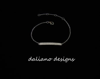 May include: A silver chain bracelet with a bar charm set with clear cubic zirconia stones. The bracelet is on a black background. "daliano designs" is written in white script below the bracelet.