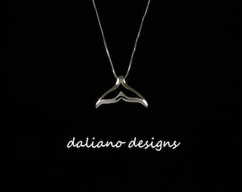 May include: A silver whale tail pendant necklace on a black background. The pendant is a simple, elegant design with a smooth, curved shape. The necklace is a delicate chain that hangs just below the collarbone. The text "daliano designs" is visible at the bottom of the image.