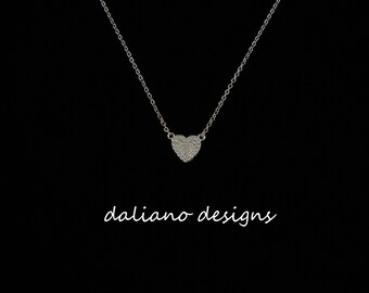 May include: A silver chain necklace with a small, heart-shaped pendant encrusted with tiny clear stones. The pendant hangs from a delicate chain. The text "daliano designs" is visible below the pendant.