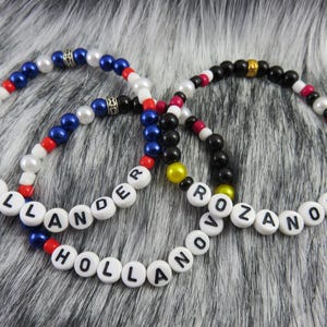 May include: Three beaded bracelets with white letter beads spelling "HOLLANDER" and "ROZANOV". The bracelets feature blue, red, white, black, and yellow beads. The bracelets are on a grey and white furry surface.