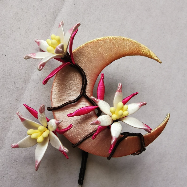 May include: A handcrafted hair accessory featuring a crescent moon shape in a soft peach color, adorned with white and pink flower accents with yellow centers. The moon is embellished with gold thread and dark brown stems.